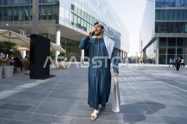 اجراء مكالمة هاتفية عبر الجوال ، الاطمئنان على الأصدقاء، تبادل الاحاديث والاخبار بين العائلة، رجل عربي خليجي اماراتي يقوم بإستخدام الأجهزة التقنية الحديثة، يحمل اكياس التسوق ، برامج وتطبيقات الجوال المتنوعة .