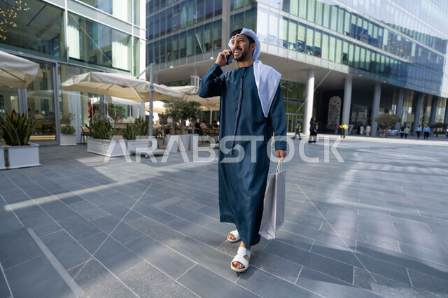 الاطمئنان على الأصدقاء، تبادل الاحاديث والاخبار بين العائلة، رجل اماراتي يقوم بإستخدام الأجهزة التقنية الحديثة، شاب عربي خليجي اماراتي مبتسم يمسك بيده جهاز الهاتف المحمول يقوم بإجراء مكالمة هاتفية ، يحمل اكياس التسوق ، برامج وتطبيقات الجوال المتنوعة .