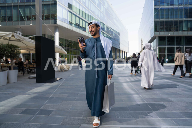 شاب عربي خليجي اماراتي مبتسم يمسك بيده جهاز الهاتف المحمول يقوم بإجراء مكالمة هاتفية ، الاطمئنان على الأصدقاء، تبادل الاحاديث والاخبار بين العائلة، رجل اماراتي يقوم بإستخدام الأجهزة التقنية الحديثة، يحمل اكياس التسوق ، برامج وتطبيقات الجوال المتنوعة .