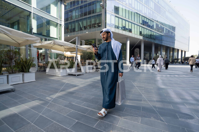 تبادل الاحاديث والاخبار بين العائلة، الاطمئنان على الأصدقاء، رجل اماراتي يقوم بإستخدام الأجهزة التقنية الحديثة، شاب عربي خليجي اماراتي مبتسم يمسك بيده جهاز الهاتف المحمول يقوم بإجراء مكالمة هاتفية ، يحمل اكياس التسوق ، برامج وتطبيقات الجوال المتنوعة .