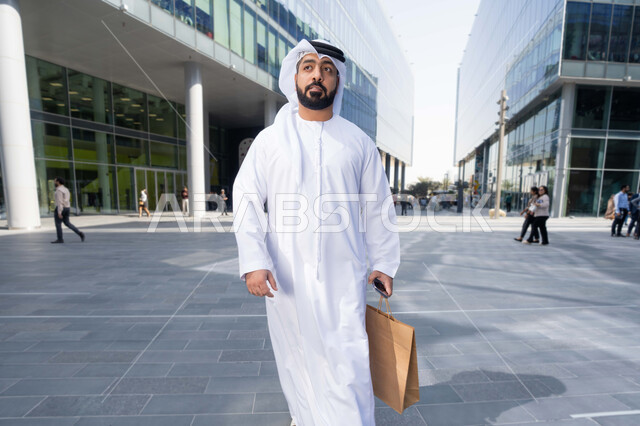التجول واستنشاق الهواء النقي في الخارج, الفخر والاعتزاز بالتقدم المعماري ، النمو والتطور العمراني في الامارات ، صورة لرجل عربي خليجي اماراتي يرتدي الكندورة والغترة البيضاء ينظر الى شيء ما في وقت النهار.