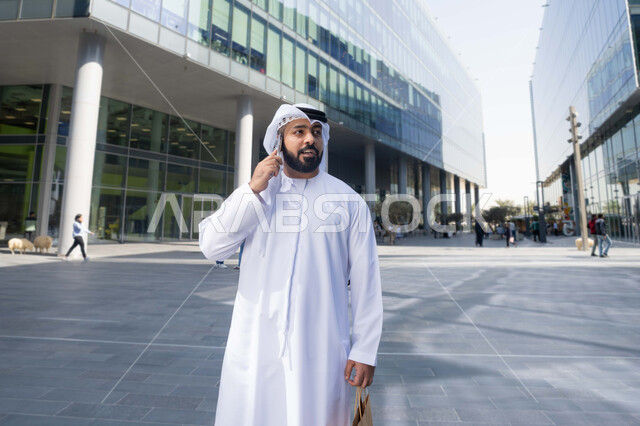 نزهة فى الهواء الطلق ، إجراء مكالمه هاتفيه عبر الهاتف المحمول، استخدام الهاتف المحمول والتقنيات الحديثه، التصفح عبر مواقع الانترنت ، رجل عربي خليجي اماراتي يرتدي الكندورة والغترة البيضاء ، ينظر بتعابير السرور يقوم بجولة فى الطبيعة ، يحمل اكياس التسوق  .
