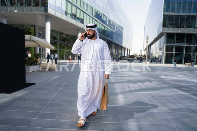 إجراء مكالمه هاتفيه عبر الهاتف المحمول، استخدام الهاتف المحمول والتقنيات الحديثه، التصفح عبر مواقع الانترنت ، رجل عربي خليجي اماراتي يرتدي الكندورة والغترة البيضاء ، ينظر بتعابير السرور يقوم بجولة فى الطبيعة ، يحمل اكياس التسوق ، نزهة فى الهواء الطلق .