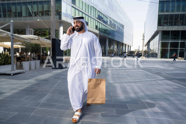 استخدام الهاتف المحمول والتقنيات الحديثه، إجراء مكالمه هاتفيه عبر الهاتف المحمول،  التصفح عبر مواقع الانترنت ، رجل عربي خليجي اماراتي يرتدي الكندورة والغترة البيضاء ، ينظر بتعابير السرور يقوم بجولة فى الطبيعة ، يحمل اكياس التسوق ، نزهة فى الهواء الطلق .