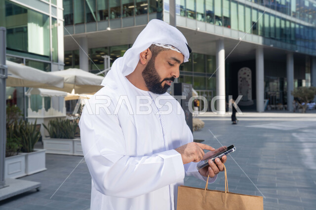 التصفح عبر مواقع الانترنت ، استخدام الهاتف المحمول والتقنيات الحديثه، الدردشة عبر مواقع التواصل الاجتماعي ، صورة مقربة لرجل عربي خليجي اماراتي يرتدي الكندورة والغترة البيضاء ، ينظر بتعابير السرور يقوم باستخدام الهاتف المحمول ، يحمل اكياس التسوق ، نزهة وجولة فى الهواء الطلق .