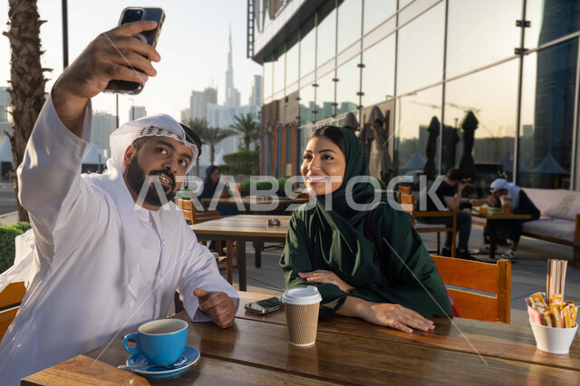 جلسات تعارف وديه, زملاء عمل في اجتماع خارج الشركة, شاب عربي خليجي اماراتي يرتدي الكندورة والغترة البيضاء يقوم باستخدام الجوال برفقة صديقته , التقاط صور سيلفي تذكارية عبر الجوال, توثيق اللحظات السعيدة, النظر إلى الكاميرا بإيماءات السعادة والسرور .