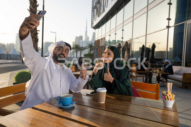 Gestures of quality and excellence, colleagues in a meeting outside the company, friendly get-to-know-you sessions, a young Emirati Gulf Arab man wearing a white kandura and ghutra using his mobile phone with his girlfriend, taking souvenir selfies with his mobile phone, documenting happy moments, looking at the camera with gestures of happiness and pleasure.