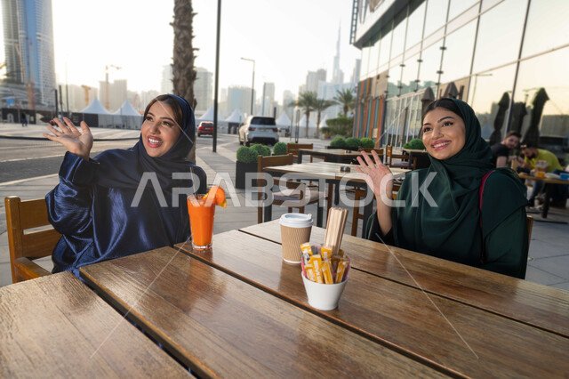 التسامر والدردشة وقضاء وقت ممتع مع الأصدقاء، جلسة وناسة يوم الاجازة، احدي المقاهي بالامارات، تطوير العلاقات الإجتماعية، تعابير السعادة والسرور بلمة الرفاق، امرأتين عربيتين خليجيتين اماراتيتين تجلسان على طاولة في المقهى، صديقتين تلقيان التحية .