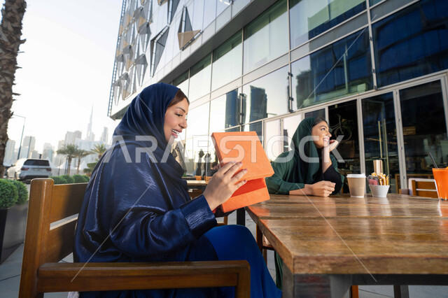 Holidays, occasions and happy surprises, a gift for Mother's Day, gestures of satisfaction and pleasure, offering gifts and flowers to mothers, the concept of mother's love and kindness towards her, a close-up photo of an Arab Gulf Emirati woman wearing an abaya sitting in a cafe, bringing a gift to her mother.