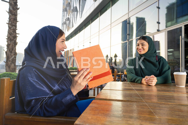 هدية بمناسبة يوم الام ، ايماءات الرضى والسرور، الاحتفال بالأعياد والمناسبات العائلية ، تقديم الهدايا والأزهار للأمهات ، مفهوم حب الأم والإحسان إليها، صورة مقربة لامراة عربية خليجية اماراتية ترتدي العباءة تجلس فى احدي المقاهي ، تقوم باحضار هدية لوالدتها .