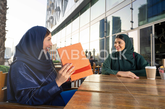 ايماءات الرضى والسرور، الاحتفال بالأعياد والمناسبات العائلية ، تقديم الهدايا والأزهار للأمهات بذكرى عيد الأم، مفهوم حب الأم والإحسان إليها، صورة مقربة لامراة عربية خليجية اماراتية ترتدي العباءة تجلس فى احدي المقاهي ، تقوم باحضار هدية لوالدتها .