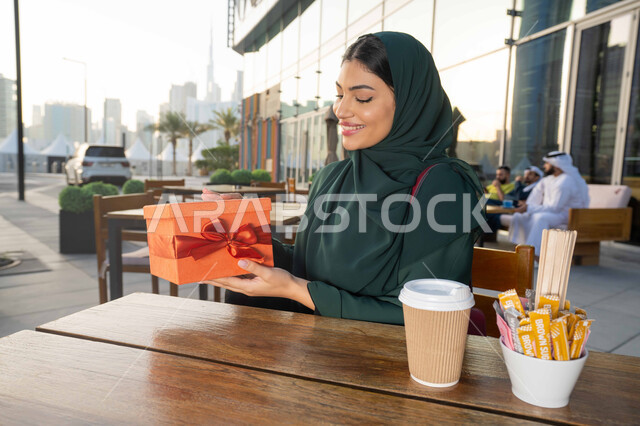 إيماءات الفرح والسعادة بالحصول على الهدايا, التعبير عن المحبة والتقدير ،الاحتفال بأعياد الميلاد, مشاركة وتقديم الهدايا في المناسبات السعيدة, صورة مقربة لامرأة عربية خليجية اماراتية ترتدي العباءة تجلس فى احدي المقاهي ، تمسك بيديها علبة هدية .