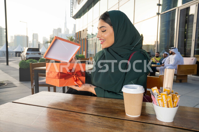 الاحتفال بأعياد الميلاد, ايماءات الدهشة والسرور, مشاركة وتقديم الهدايا في المناسبات السعيدة, صورة مقربة لامرأة عربية خليجية اماراتية ترتدي العباءة تجلس فى احدي المقاهي ، تمسك بيديها علبة هدية, التعبير عن المحبة والتقدير .