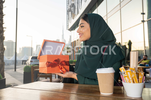 التعبير عن المحبة والتقدير ،الاحتفال بأعياد الميلاد, ايماءات الدهشة والسرور, مشاركة وتقديم الهدايا في المناسبات السعيدة, صورة مقربة لامرأة عربية خليجية اماراتية ترتدي العباءة تجلس فى احدي المقاهي ، تمسك بيديها علبة هدية .