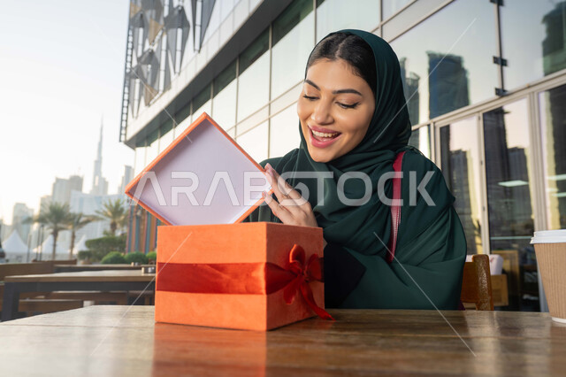 ايماءات الدهشة والسرور, الاحتفال بأعياد الميلاد, مشاركة وتقديم الهدايا في المناسبات السعيدة, صورة مقربة لامرأة عربية خليجية اماراتية ترتدي العباءة تجلس فى احدي المقاهي ، تمسك بيديها علبة هدية, التعبير عن المحبة والتقدير .