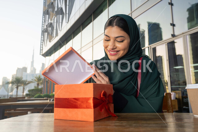 مشاركة وتقديم الهدايا في المناسبات السعيدة, ايماءات الدهشة والسرور, الاحتفال بأعياد الميلاد, صورة مقربة لامرأة عربية خليجية اماراتية ترتدي العباءة تجلس فى احدي المقاهي ، تمسك بيديها علبة هدية, التعبير عن المحبة والتقدير .