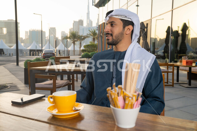 الاستمتاع باجواء هادئه خارج المنزل ، رجل عربي اماراتي خليجي يرتدي الكندورة والغترة البيضاء يجلس في المقهى, الاستمتاع بشرب القهوه, انجاز بعض مهام العمل في المقهى, العمل عن بعد, بناء علاقات وصداقات جديدة , انجاز المهام اليوميه .