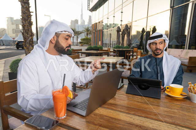تصفح مواقع التواصل الإجتماعي والعمل عن بعد، الحوار والنقاش بين زملاء العمل ، ايماءات الاندماج والتركيز ، قضاء وقت ممتع مع الأصدقاء, استخدم جهاز الحاسوب المحمول لانجاز المشاريع والمهام في الكافيه، صورة مقربة لشاب عربي خليجي إماراتي يرتدي الكندورة والغترة البيضاء يجلس مع صديقه في المقهى  .