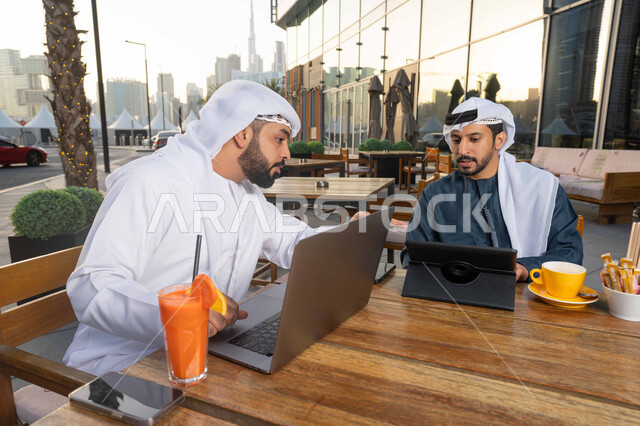 الحوار والنقاش بين زملاء العمل ، ايماءات الاندماج والتركيز ، قضاء وقت ممتع مع الأصدقاء, استخدم جهاز الحاسوب المحمول لانجاز المشاريع والمهام في الكافيه، صورة مقربة لشاب عربي خليجي إماراتي يرتدي الكندورة والغترة البيضاء يجلس مع صديقه في المقهى  .