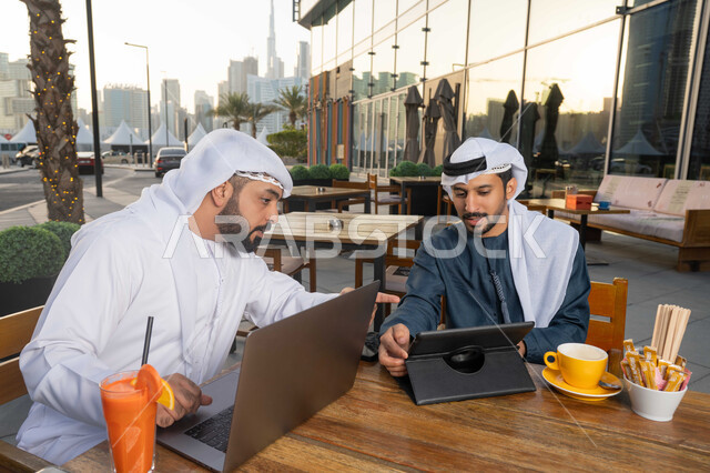 قضاء وقت ممتع مع الأصدقاء, استخدم جهاز الحاسوب المحمول لانجاز المشاريع والمهام في الكافيه، الحوار والنقاش بين زملاء العمل، صورة مقربة لشاب عربي خليجي إماراتي يرتدي الكندورة والغترة البيضاء يجلس مع صديقه في المقهى ، ايماءات الاندماج والتركيز .