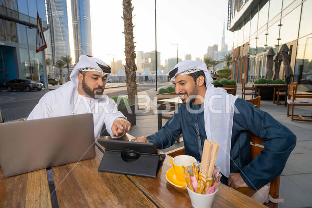 Using the laptop to complete projects and tasks in the cafe, dialogue and discussion between colleagues, spending quality time with friends, a close-up of an Emirati Gulf Arab young man wearing a white kandura and ghutra sitting with his friend in the cafe, gestures of integration and concentration.