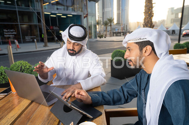 الحوار والنقاش بين زملاء العمل، استخدم جهاز الحاسوب المحمول لانجاز المشاريع والمهام في الكافيه، قضاء وقت ممتع مع الأصدقاء, صورة مقربة لشاب عربي خليجي إماراتي يرتدي الكندورة والغترة البيضاء يجلس مع صديقه في المقهى ، ايماءات الاندماج والتركيز .