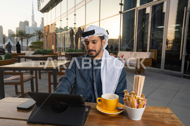 إدارة شؤون الأعمال وتنظيمها عبر الأجهزة التقنية الحديثة، استخدام التكنولوجيا والتقنيات المتطورة، إنجاز مهام العمل عن بعد،  صورة لرجل عربي خليجي اماراتي يرتدي الكندورة والغترة البيضاء يجلس على كرسي في إحدى المقاهي يشرب مشروب ساخن ويستخدم الحاسوب المحمول .