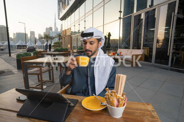 Using advanced technology and techniques, completing work tasks remotely, managing and organizing business affairs through modern technical devices, a picture of an Emirati Gulf Arab man wearing a white kandura and ghutra sitting on a chair in a cafe drinking a hot drink and using a laptop.