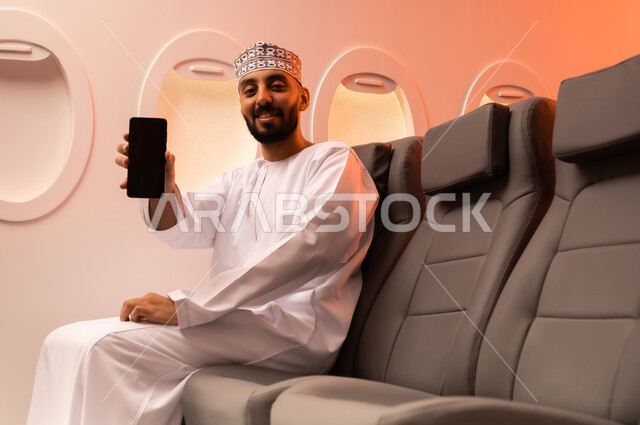 الترفيه والتسليه في اوقات الفراغ,  تصفح وسائل التواصل الاجتماعي, استخدام جهاز تقني حديث ومتطور, بورتريه مقرب من الجانب لرجل عربي خليجي عماني يرتدي الدشداشة والكمة يمسك بيده الهاتف المحمول جالسا على كرسيه داخل الطائره الخليجيه, الطيران الخليجي العماني