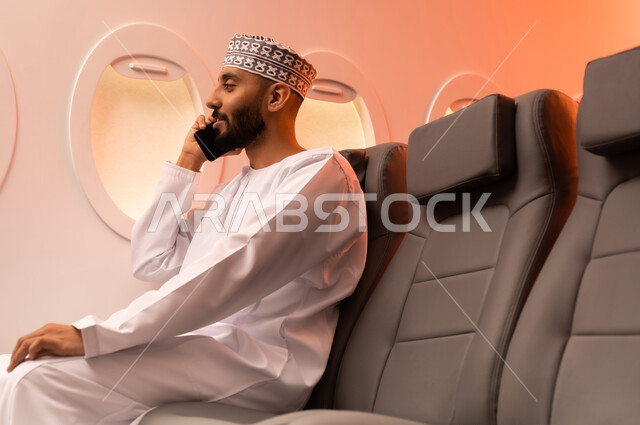 الترفيه والتسليه في اوقات الفراغ,  تصفح وسائل التواصل الاجتماعي, استخدام جهاز تقني حديث ومتطور, بورتريه مقرب من الجانب لرجل عربي خليجي عماني يرتدي الدشداشة والكمة يمسك بيده الهاتف المحمول جالسا على كرسيه داخل الطائره الخليجيه, الطيران الخليجي العماني
