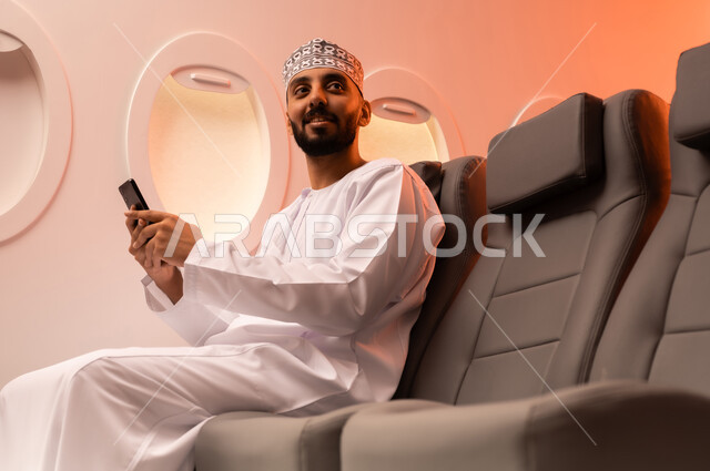الترفيه والتسليه في اوقات الفراغ,  تصفح وسائل التواصل الاجتماعي, استخدام جهاز تقني حديث ومتطور, بورتريه مقرب من الجانب لرجل عربي خليجي عماني يرتدي الدشداشة والكمة يمسك بيده الهاتف المحمول جالسا على كرسيه داخل الطائره الخليجيه, الطيران الخليجي العماني