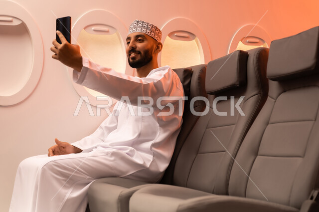الترفيه والتسليه في اوقات الفراغ,  تصفح وسائل التواصل الاجتماعي, استخدام جهاز تقني حديث ومتطور, بورتريه مقرب من الجانب لرجل عربي خليجي عماني يرتدي الدشداشة والكمة يمسك بيده الهاتف المحمول جالسا على كرسيه داخل الطائره الخليجيه, الطيران الخليجي العماني