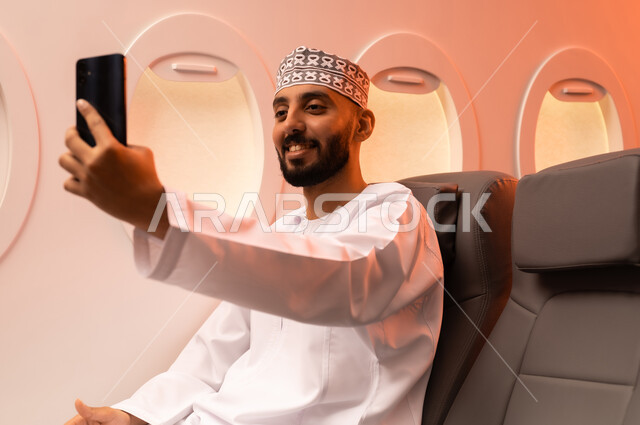 الترفيه والتسليه في اوقات الفراغ,  تصفح وسائل التواصل الاجتماعي, استخدام جهاز تقني حديث ومتطور, بورتريه مقرب من الجانب لرجل عربي خليجي عماني يرتدي الدشداشة والكمة يمسك بيده الهاتف المحمول جالسا على كرسيه داخل الطائره الخليجيه, الطيران الخليجي العماني