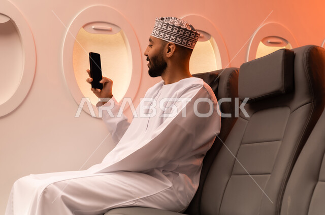 الترفيه والتسليه في اوقات الفراغ,  تصفح وسائل التواصل الاجتماعي, استخدام جهاز تقني حديث ومتطور, بورتريه مقرب من الجانب لرجل عربي خليجي عماني يرتدي الدشداشة والكمة يمسك بيده الهاتف المحمول جالسا على كرسيه داخل الطائره الخليجيه, الطيران الخليجي العماني