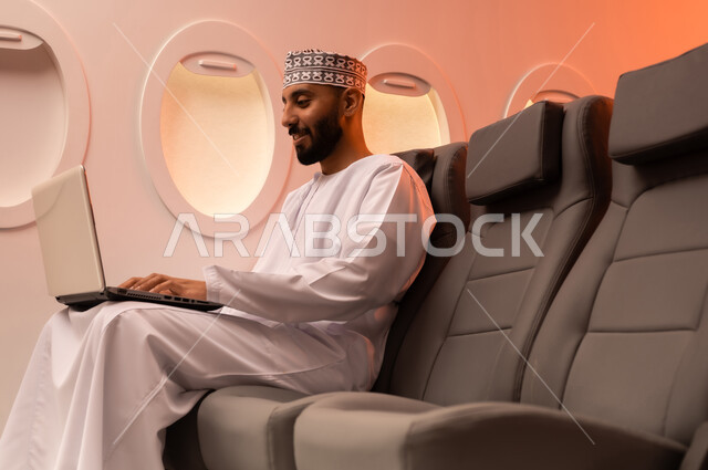 مفهوم ممارسه الانشطه والعمل عن بعد داخل الطائره, بورتريه مقرب من الجانب  لرجل عربي خليجي عماني يرتدي الدشداشه والكمه يجلس على كرسيه داخل الطائره الخليجيه يقوم بالعمل على جهاز الحاسوب المحمول, الطيران العماني الخليجي
