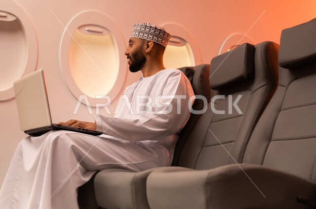 مفهوم ممارسه الانشطه والعمل عن بعد داخل الطائره, بورتريه مقرب من الجانب  لرجل عربي خليجي عماني يرتدي الدشداشه والكمه يجلس على كرسيه داخل الطائره الخليجيه يقوم بالعمل على جهاز الحاسوب المحمول, الطيران العماني الخليجي