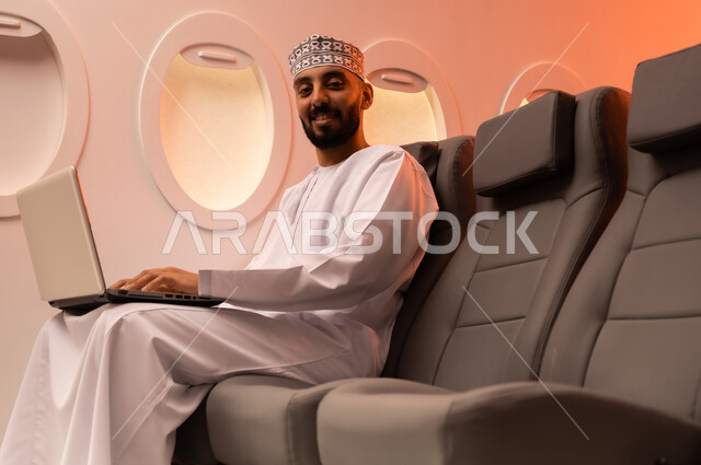 مفهوم ممارسه الانشطه والعمل عن بعد داخل الطائره, بورتريه مقرب من الجانب  لرجل عربي خليجي عماني يرتدي الدشداشه والكمه يجلس على كرسيه داخل الطائره الخليجيه يقوم بالعمل على جهاز الحاسوب المحمول, الطيران العماني الخليجي