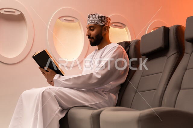 ممارسه الانشطه الترفيهيه وهوايه المطالعه, بورتريه مقرب من الجانب  لرجل عربي خليجي عماني يرتدي الدشداشه والكمه يجلس على كرسيه داخل الطائره يقرأ كتابا بيده, الطيران العماني الخليجي