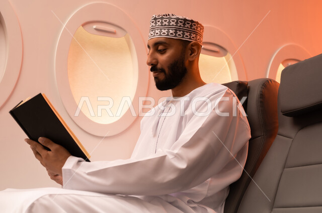 ممارسه الانشطه الترفيهيه وهوايه المطالعه, بورتريه مقرب من الجانب  لرجل عربي خليجي عماني يرتدي الدشداشه والكمه يجلس على كرسيه داخل الطائره يقرأ كتابا بيده, الطيران العماني الخليجي