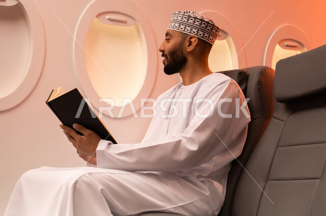 ممارسه الانشطه الترفيهيه وهوايه المطالعه, بورتريه مقرب من الجانب  لرجل عربي خليجي عماني يرتدي الدشداشه والكمه يجلس على كرسيه داخل الطائره يقرأ كتابا بيده, الطيران العماني الخليجي