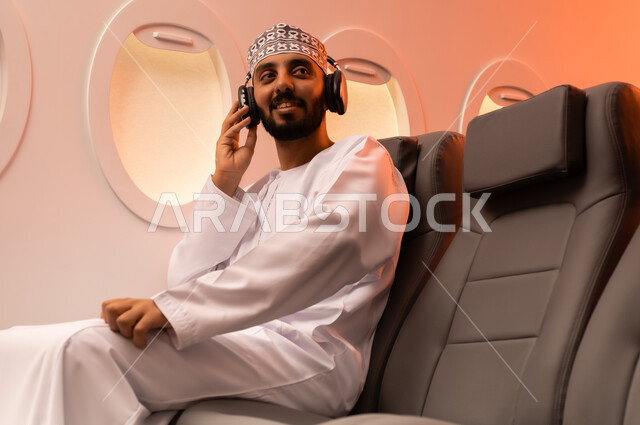 استخدام تقنية الهيدفون، الاستماع الى الأغاني والموسيقى عبر سماعة الرأس، بورتريه مقرب من الجانب  لرجل عربي خليجي عماني يرتدي الدشداشه والكمه يجلس على كرسيه داخل الطائره يضع سماعة الرأس ويقوم بالإصغاء والاستماع إلى شيء ما، طيران عمان