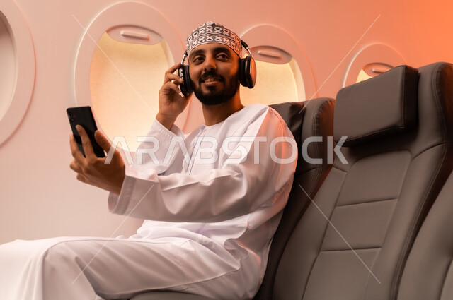 استخدام تقنية الهيدفون، الاستماع الى الأغاني والموسيقى عبر سماعة الرأس، بورتريه مقرب من الجانب  لرجل عربي خليجي عماني يرتدي الدشداشه والكمه يجلس على كرسيه داخل الطائره يضع سماعة الرأس ويقوم بالإصغاء والاستماع إلى شيء ما، طيران عمان