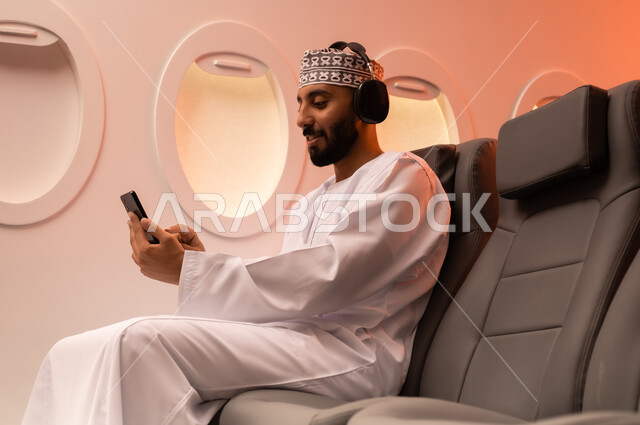 استخدام تقنية الهيدفون، الاستماع الى الأغاني والموسيقى عبر سماعة الرأس، بورتريه مقرب من الجانب  لرجل عربي خليجي عماني يرتدي الدشداشه والكمه يجلس على كرسيه داخل الطائره يضع سماعة الرأس ويقوم بالإصغاء والاستماع إلى شيء ما، طيران عمان