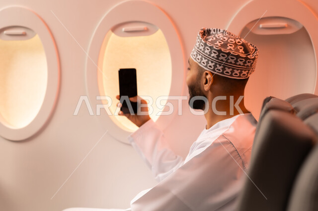 توثيق اللحظات التذكارية عبر الهاتف المحمول,  السياحة في إجازة الصيف, بورتريه مقرب من الجانب لشاب عربي خليجي عماني يرتدي الدشداشه والكمه يجلس على كرسيه داخل الطائره ويمسك بيده هاتفهه المحمول يلتقط صور سيلفي استعدادا  لقضاء رحلة صيفية ممتعة