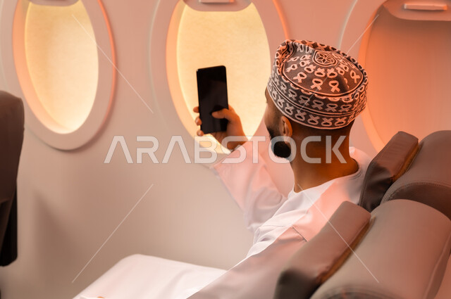 توثيق اللحظات التذكارية عبر الهاتف المحمول,  السياحة في إجازة الصيف, بورتريه مقرب من الجانب لشاب عربي خليجي عماني يرتدي الدشداشه والكمه يجلس على كرسيه داخل الطائره ويمسك بيده هاتفهه المحمول يلتقط صور سيلفي استعدادا  لقضاء رحلة صيفية ممتعة