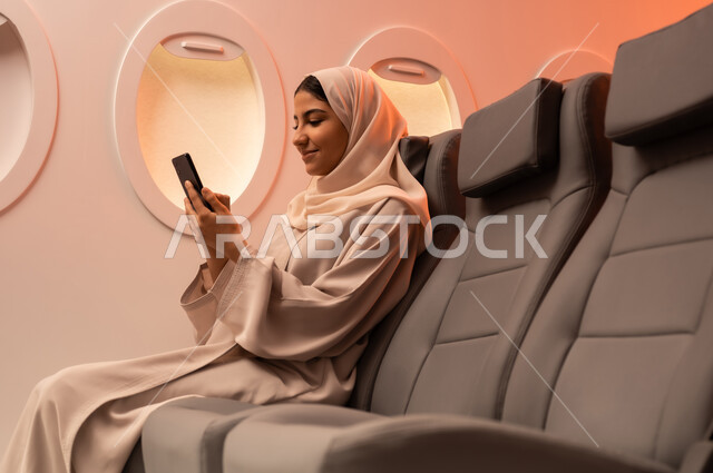 Documenting memorable moments via mobile phone, summer vacation tourism, close-up side portrait of a young Arab Gulf Omani woman wearing an abaya and hijab sitting on her seat inside the plane and holding her mobile phone in her hand, preparing for a pleasant summer trip