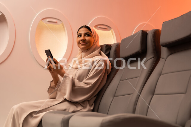 توثيق اللحظات التذكارية عبر الهاتف المحمول,  السياحة في إجازة الصيف, بورتريه مقرب من الجانب لشابه عربيه خليجيه عمانيه ترتدي العبائه والحجاب تجلس على كرسيها داخل الطائره وتمسك بيدها هاتفهها المحمول تستعد لقضاء رحلة صيفية ممتعة