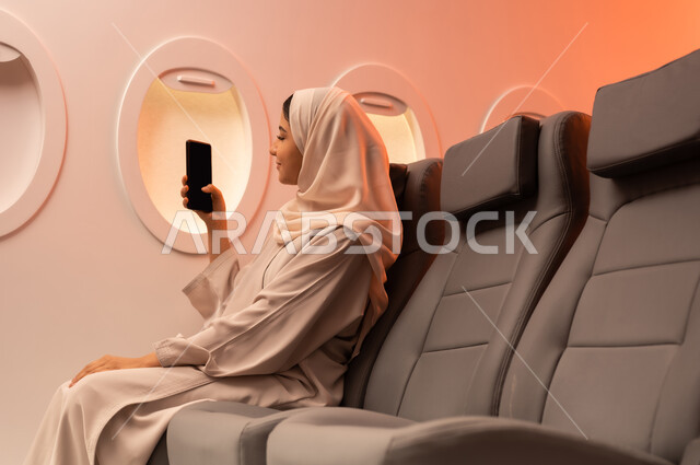Documenting memorable moments via mobile phone, summer vacation tourism, close-up side portrait of a young Arab Gulf Omani woman wearing an abaya and hijab sitting on her seat inside the plane and holding her mobile phone in her hand, preparing for a pleasant summer trip