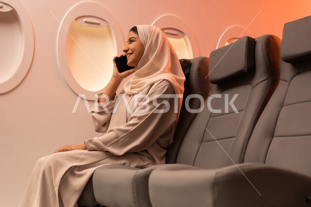 توثيق اللحظات التذكارية عبر الهاتف المحمول,  السياحة في إجازة الصيف, بورتريه مقرب من الجانب لشابه عربيه خليجيه عمانيه ترتدي العبائه والحجاب تجلس على كرسيها داخل الطائره وتمسك بيدها هاتفهها المحمول تستعد لقضاء رحلة صيفية ممتعة