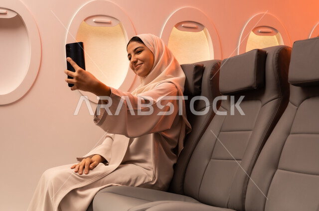 توثيق اللحظات التذكارية عبر الهاتف المحمول,  السياحة في إجازة الصيف, بورتريه مقرب من الجانب لشابه عربيه خليجيه عمانيه ترتدي العبائه والحجاب تجلس على كرسيها داخل الطائره وتمسك بيدها هاتفهها المحمول تستعد لقضاء رحلة صيفية ممتعة